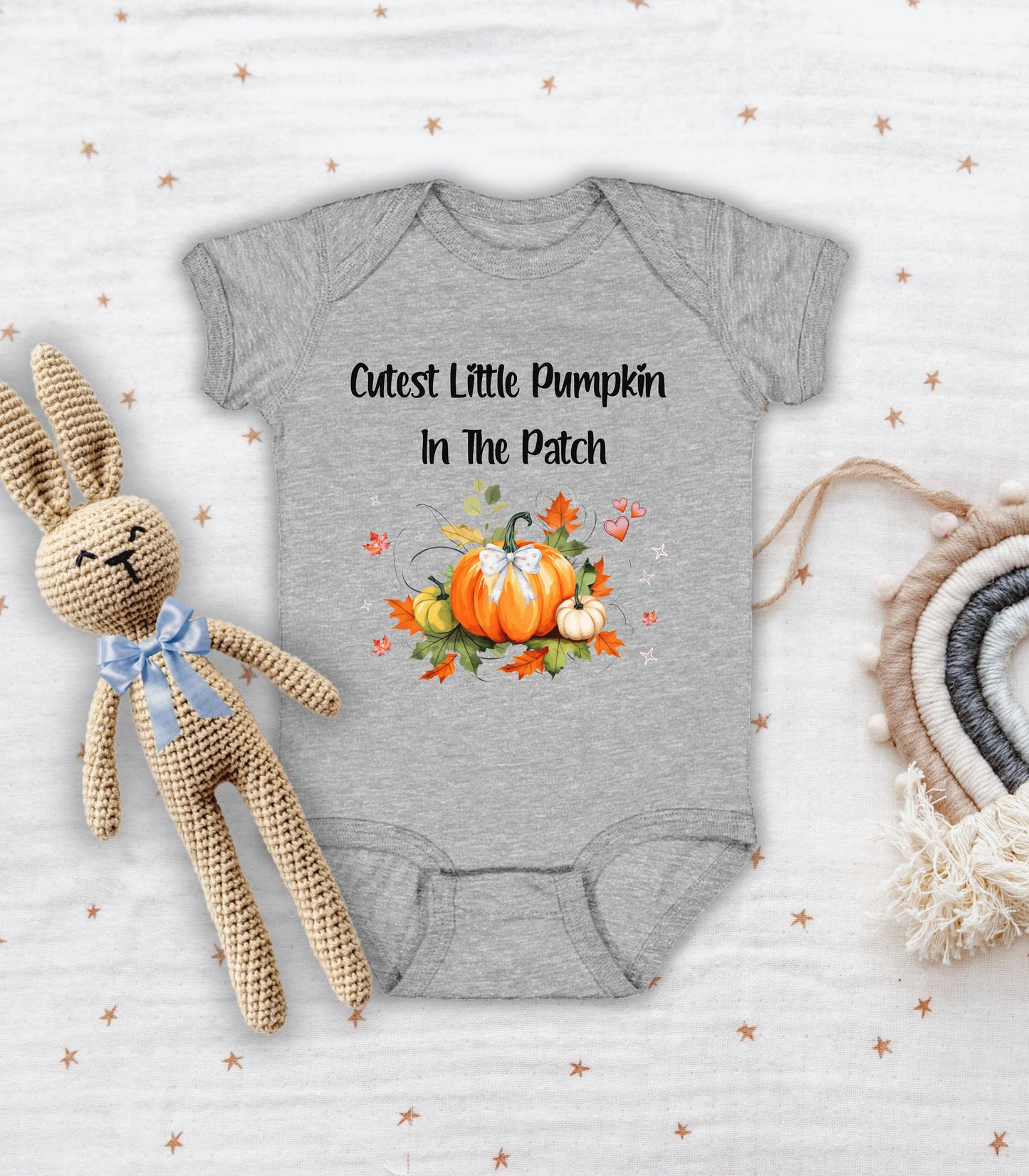 Heather infant bodysuit featuring the phrase "Cutest Little Pumpkin In The Patch" with an adorable fall-themed design of orange and white pumpkins, autumn leaves, hearts, and a sweet bow. Perfect for baby's first fall or pumpkin patch visit.