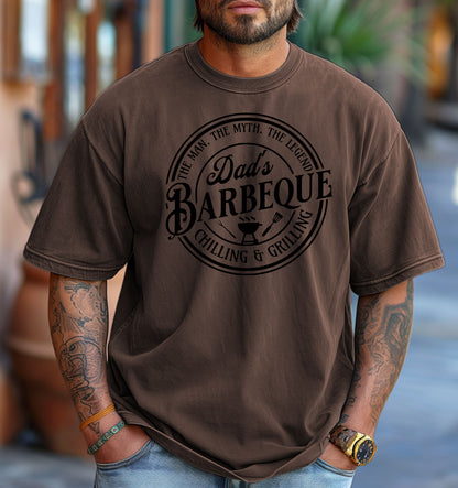 Dad’s Barbeque T-shirt featuring a circular badge-style design with the phrases 'The Man. The Myth. The Legend.' and 'Chilling & Grilling.' The center showcases 'Dad’s Barbeque' in bold lettering with a small grill, fork, and spatula icons. Printed on a espresso unisex tee, this shirt is perfect for BBQ-loving dads and makes a fun Father’s Day or birthday gift.