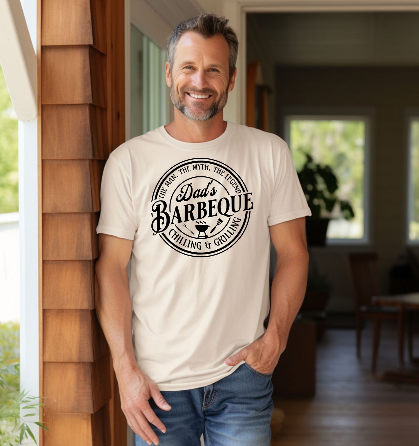 Dad’s Barbeque T-shirt featuring a circular badge-style design with the phrases 'The Man. The Myth. The Legend.' and 'Chilling & Grilling.' The center showcases 'Dad’s Barbeque' in bold lettering with a small grill, fork, and spatula icons. Printed on an ivory unisex tee, this shirt is perfect for BBQ-loving dads and makes a fun Father’s Day or birthday gift.