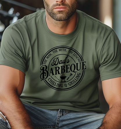 Dad’s Barbeque T-shirt featuring a circular badge-style design with the phrases 'The Man. The Myth. The Legend.' and 'Chilling & Grilling.' The center showcases 'Dad’s Barbeque' in bold lettering with a small grill, fork, and spatula icons. Printed on a moss unisex tee, this shirt is perfect for BBQ-loving dads and makes a fun Father’s Day or birthday gift.