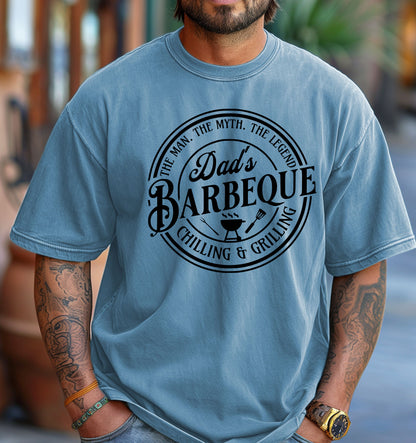 Dad’s Barbeque T-shirt featuring a circular badge-style design with the phrases 'The Man. The Myth. The Legend.' and 'Chilling & Grilling.' The center showcases 'Dad’s Barbeque' in bold lettering with a small grill, fork, and spatula icons. Printed on a washed denim unisex tee, this shirt is perfect for BBQ-loving dads and makes a fun Father’s Day or birthday gift.