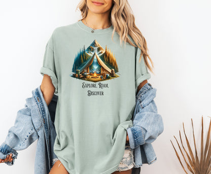Bay “Explore, Roam, Discover” T-shirt featuring a scenic camping design with an open tent framing a mountain, waterfall, pine forest, and river. A cozy campfire, gear, and canoe paddles sit in the foreground, creating an adventurous and tranquil outdoor vibe. Perfect for nature lovers, hikers, and camping enthusiasts seeking wanderlust-inspired apparel.