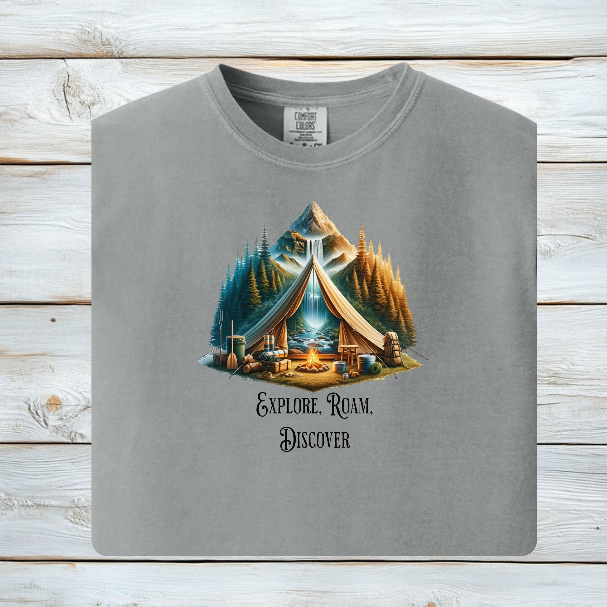 Granite “Explore, Roam, Discover” T-shirt featuring a scenic camping design with an open tent framing a mountain, waterfall, pine forest, and river. A cozy campfire, gear, and canoe paddles sit in the foreground, creating an adventurous and tranquil outdoor vibe. Perfect for nature lovers, hikers, and camping enthusiasts seeking wanderlust-inspired apparel.