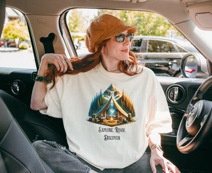 Ivory “Explore, Roam, Discover” T-shirt featuring a scenic camping design with an open tent framing a mountain, waterfall, pine forest, and river. A cozy campfire, gear, and canoe paddles sit in the foreground, creating an adventurous and tranquil outdoor vibe. Perfect for nature lovers, hikers, and camping enthusiasts seeking wanderlust-inspired apparel.