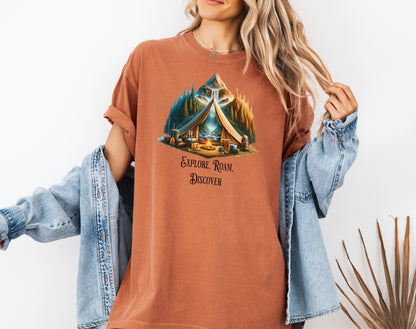 Yam “Explore, Roam, Discover” T-shirt featuring a scenic camping design with an open tent framing a mountain, waterfall, pine forest, and river. A cozy campfire, gear, and canoe paddles sit in the foreground, creating an adventurous and tranquil outdoor vibe. Perfect for nature lovers, hikers, and camping enthusiasts seeking wanderlust-inspired apparel.