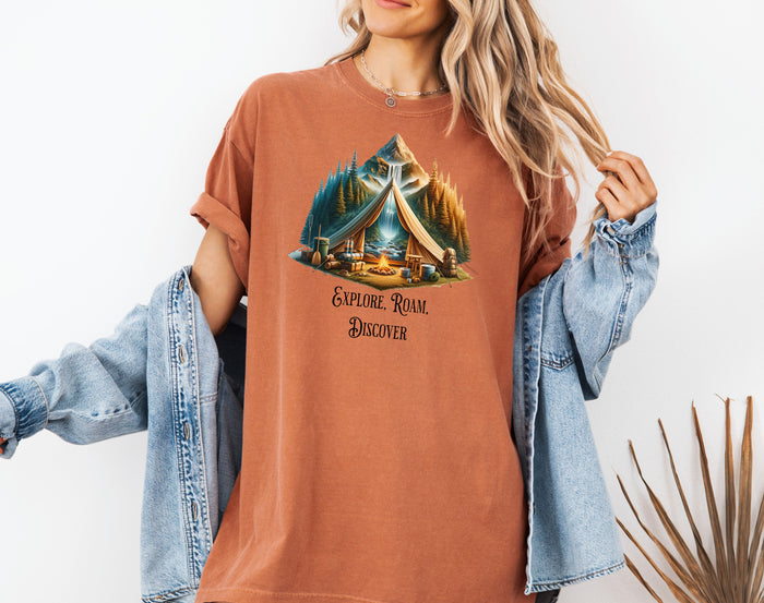 Yam “Explore, Roam, Discover” T-shirt featuring a scenic camping design with an open tent framing a mountain, waterfall, pine forest, and river. A cozy campfire, gear, and canoe paddles sit in the foreground, creating an adventurous and tranquil outdoor vibe. Perfect for nature lovers, hikers, and camping enthusiasts seeking wanderlust-inspired apparel.