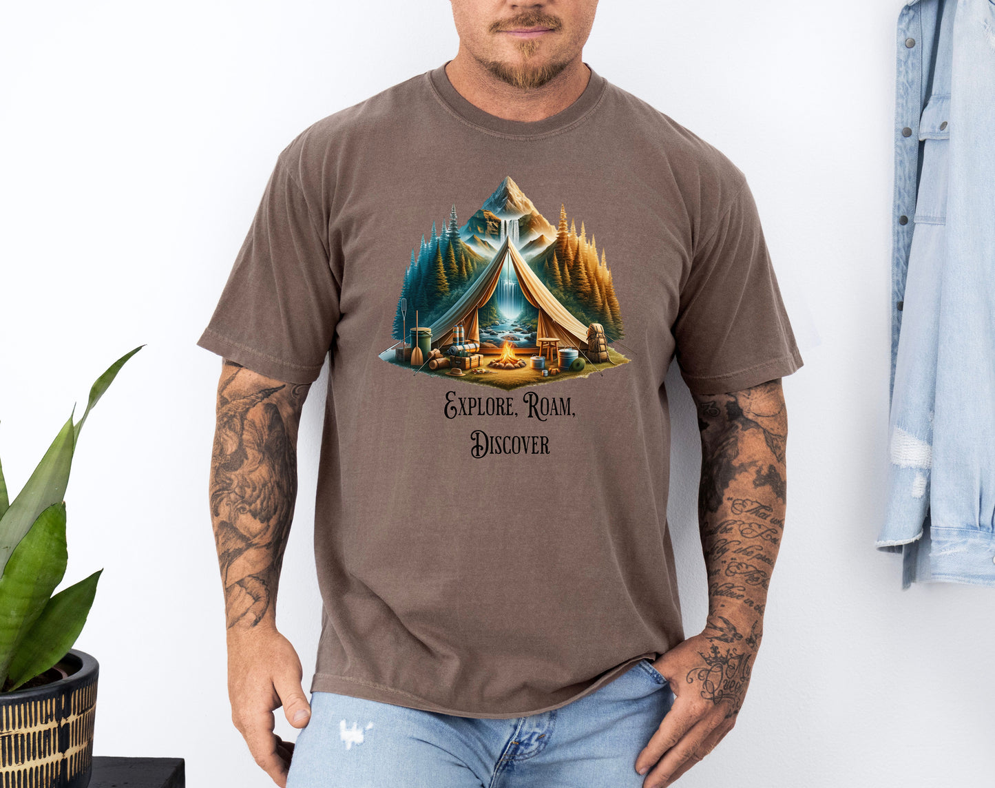 Espresso “Explore, Roam, Discover” T-shirt featuring a scenic camping design with an open tent framing a mountain, waterfall, pine forest, and river. A cozy campfire, gear, and canoe paddles sit in the foreground, creating an adventurous and tranquil outdoor vibe. Perfect for nature lovers, hikers, and camping enthusiasts seeking wanderlust-inspired apparel.