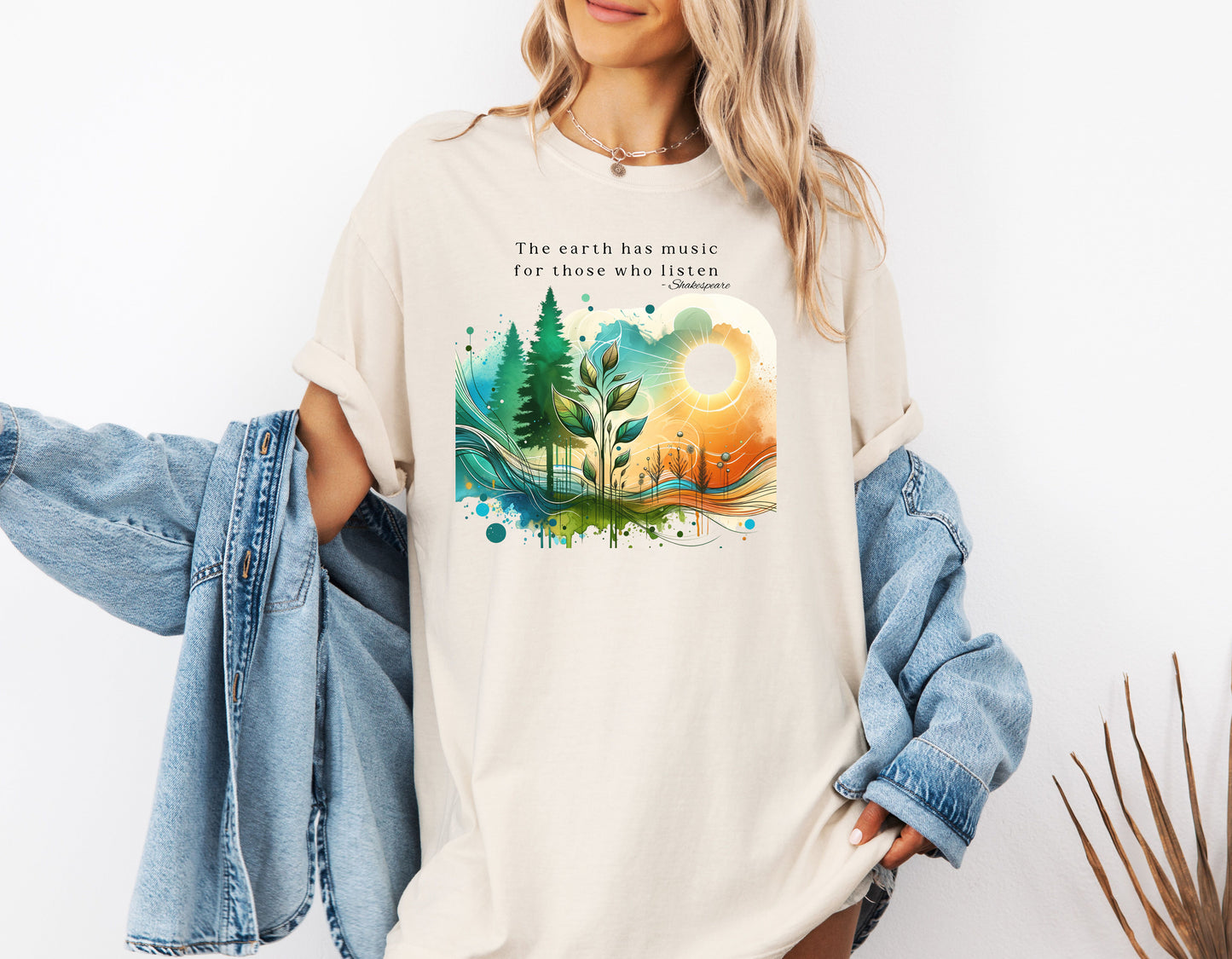 An Ivory T-shirt featuring a vibrant “Earth Music” nature design with the quote “The earth has music for those who listen – Shakespeare.” Artwork showcases a sunrise over green pine trees, flowing waves, and a sprouting plant, blending watercolor textures with bright greens, blues, and oranges.  This eco-inspired graphic tee celebrates the beauty and harmony of nature.
