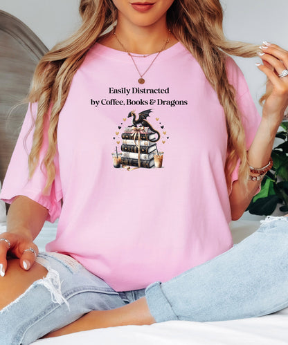 Blossom pink t-shirt featuring the phrase “Easily Distracted by Coffee, Books & Dragons” black text above a whimsical design of a black dragon perched on a stack of ornate books, surrounded by gold hearts and iced coffee cups. A fun and magical design perfect for fantasy lovers who adore caffeine and getting lost in a good book.