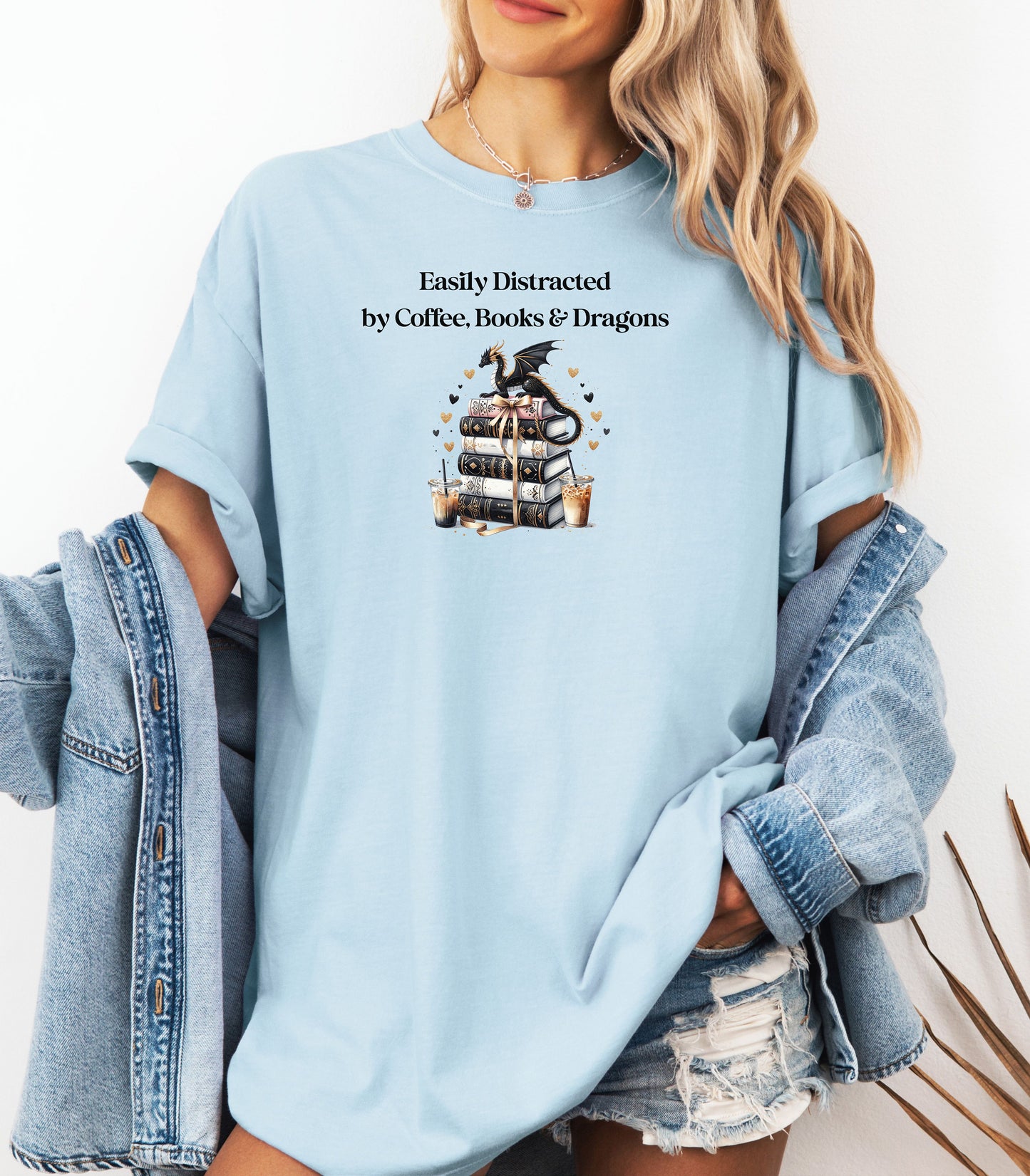 Chambray light blue t-shirt featuring the phrase “Easily Distracted by Coffee, Books & Dragons” black text above a whimsical design of a black dragon perched on a stack of ornate books, surrounded by gold hearts and iced coffee cups. A fun and magical design perfect for fantasy lovers who adore caffeine and getting lost in a good book.