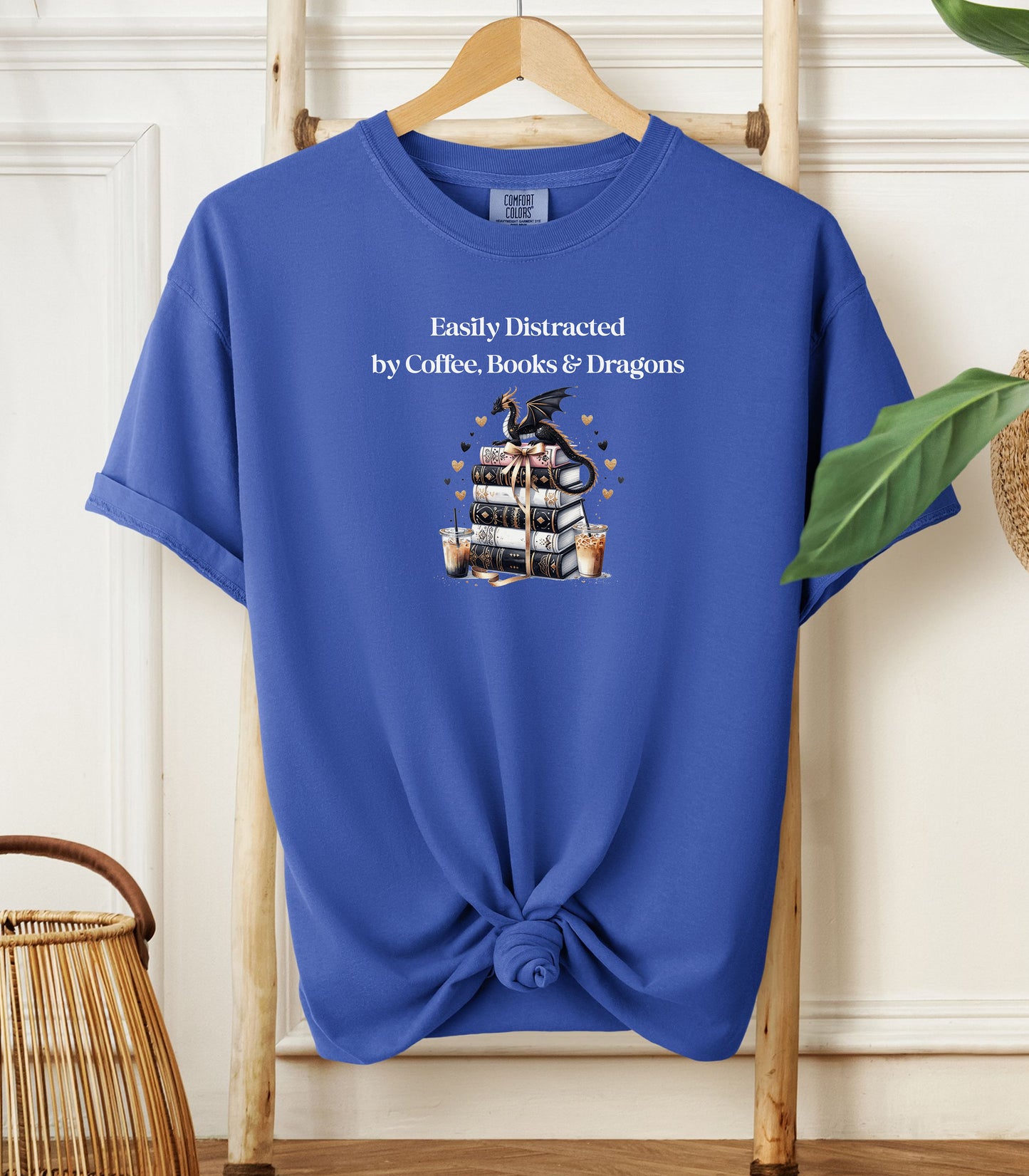 Flo blue t-shirt featuring the phrase “Easily Distracted by Coffee, Books & Dragons” white text above a whimsical design of a black dragon perched on a stack of ornate books, surrounded by gold hearts and iced coffee cups. A fun and magical design perfect for fantasy lovers who adore caffeine and getting lost in a good book.