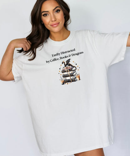 White t-shirt featuring the phrase “Easily Distracted by Coffee, Books & Dragons” black text above a whimsical design of a black dragon perched on a stack of ornate books, surrounded by gold hearts and iced coffee cups. A fun and magical design perfect for fantasy lovers who adore caffeine and getting lost in a good book.