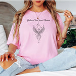 Person wearing a blossom pink T-shirt featuring an elegant black line art phoenix design with wings spread wide, flowing tail feathers, and celestial elements including a moon and stars. Above the artwork, the phrase “Embrace Your Inner Phoenix” is printed in a graceful script font, symbolizing strength, rebirth, and transformation. Perfect for those who value empowerment, resilience, and a bold yet feminine style.