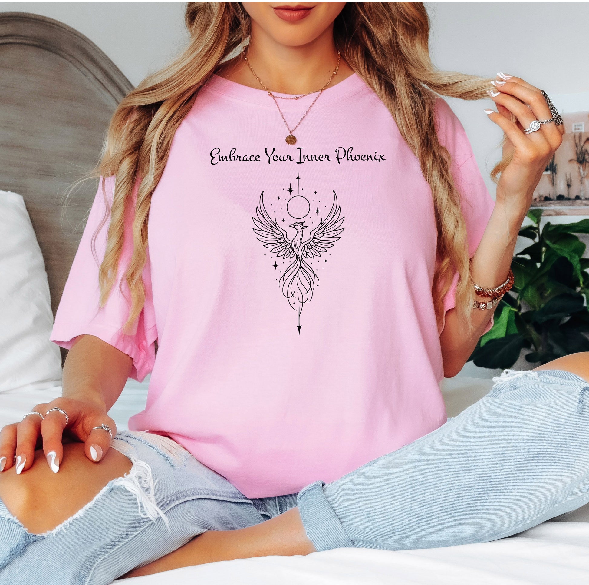 Person wearing a blossom pink T-shirt featuring an elegant black line art phoenix design with wings spread wide, flowing tail feathers, and celestial elements including a moon and stars. Above the artwork, the phrase “Embrace Your Inner Phoenix” is printed in a graceful script font, symbolizing strength, rebirth, and transformation. Perfect for those who value empowerment, resilience, and a bold yet feminine style.