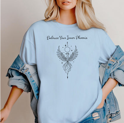 Person wearing a chambray light blue T-shirt featuring an elegant black line art phoenix design with wings spread wide, flowing tail feathers, and celestial elements including a moon and stars. Above the artwork, the phrase “Embrace Your Inner Phoenix” is printed in a graceful script font, symbolizing strength, rebirth, and transformation. Perfect for those who value empowerment, resilience, and a bold yet feminine style.