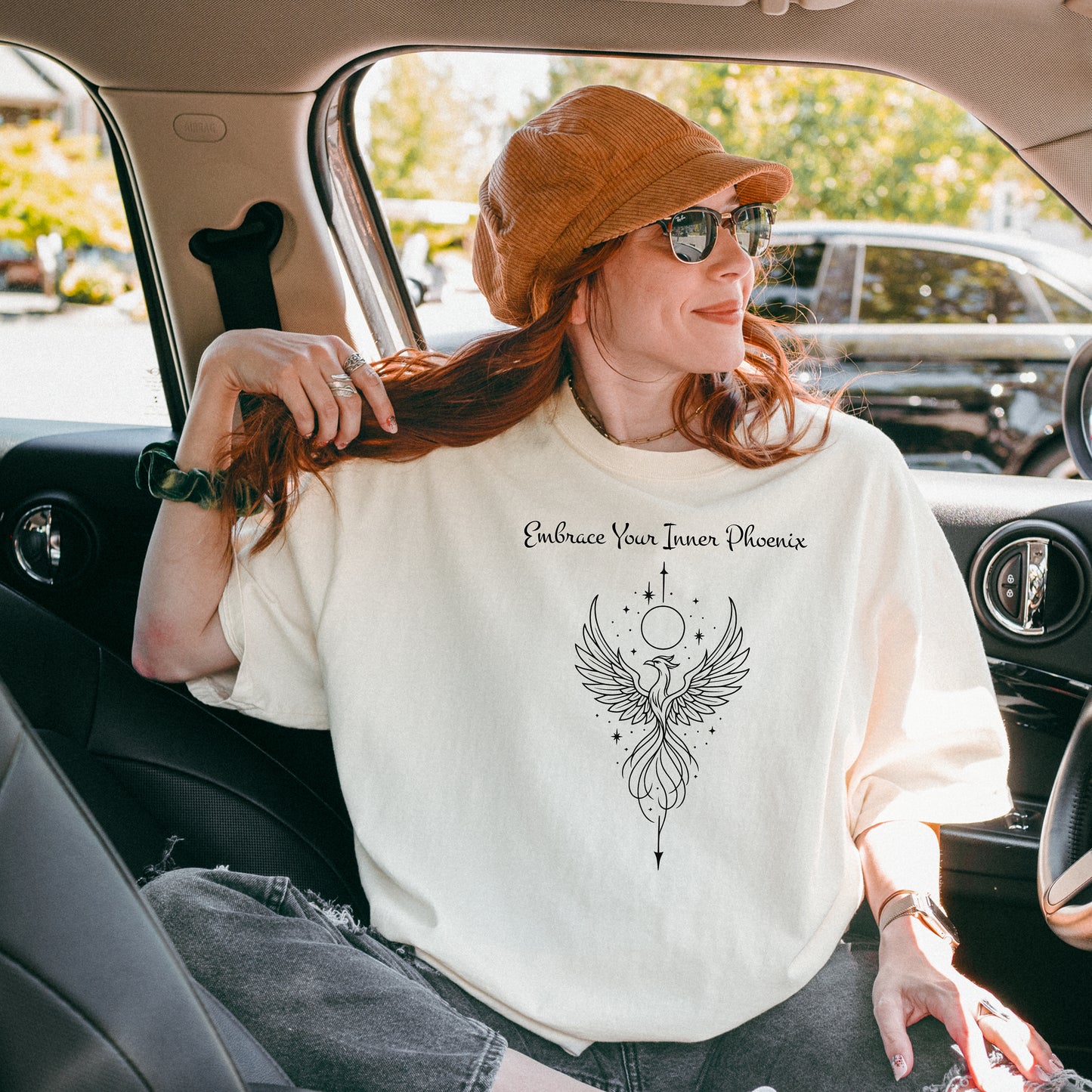 Person wearing an ivory T-shirt featuring an elegant black line art phoenix design with wings spread wide, flowing tail feathers, and celestial elements including a moon and stars. Above the artwork, the phrase “Embrace Your Inner Phoenix” is printed in a graceful script font, symbolizing strength, rebirth, and transformation. Perfect for those who value empowerment, resilience, and a bold yet feminine style.