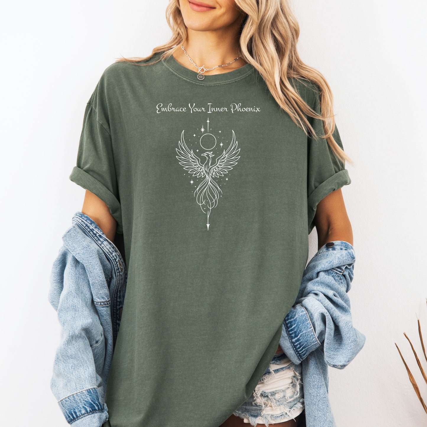 Person wearing a moss T-shirt featuring an elegant black line art phoenix design with wings spread wide, flowing tail feathers, and celestial elements including a moon and stars. Above the artwork, the phrase “Embrace Your Inner Phoenix” is printed in a graceful script font, symbolizing strength, rebirth, and transformation. Perfect for those who value empowerment, resilience, and a bold yet feminine style.