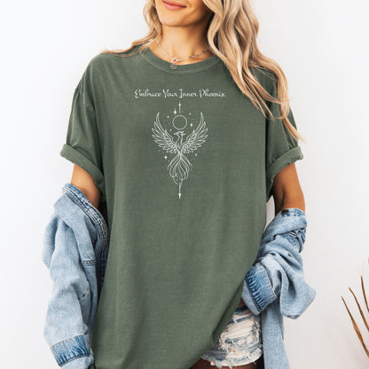 Person wearing a moss T-shirt featuring an elegant black line art phoenix design with wings spread wide, flowing tail feathers, and celestial elements including a moon and stars. Above the artwork, the phrase “Embrace Your Inner Phoenix” is printed in a graceful script font, symbolizing strength, rebirth, and transformation. Perfect for those who value empowerment, resilience, and a bold yet feminine style.
