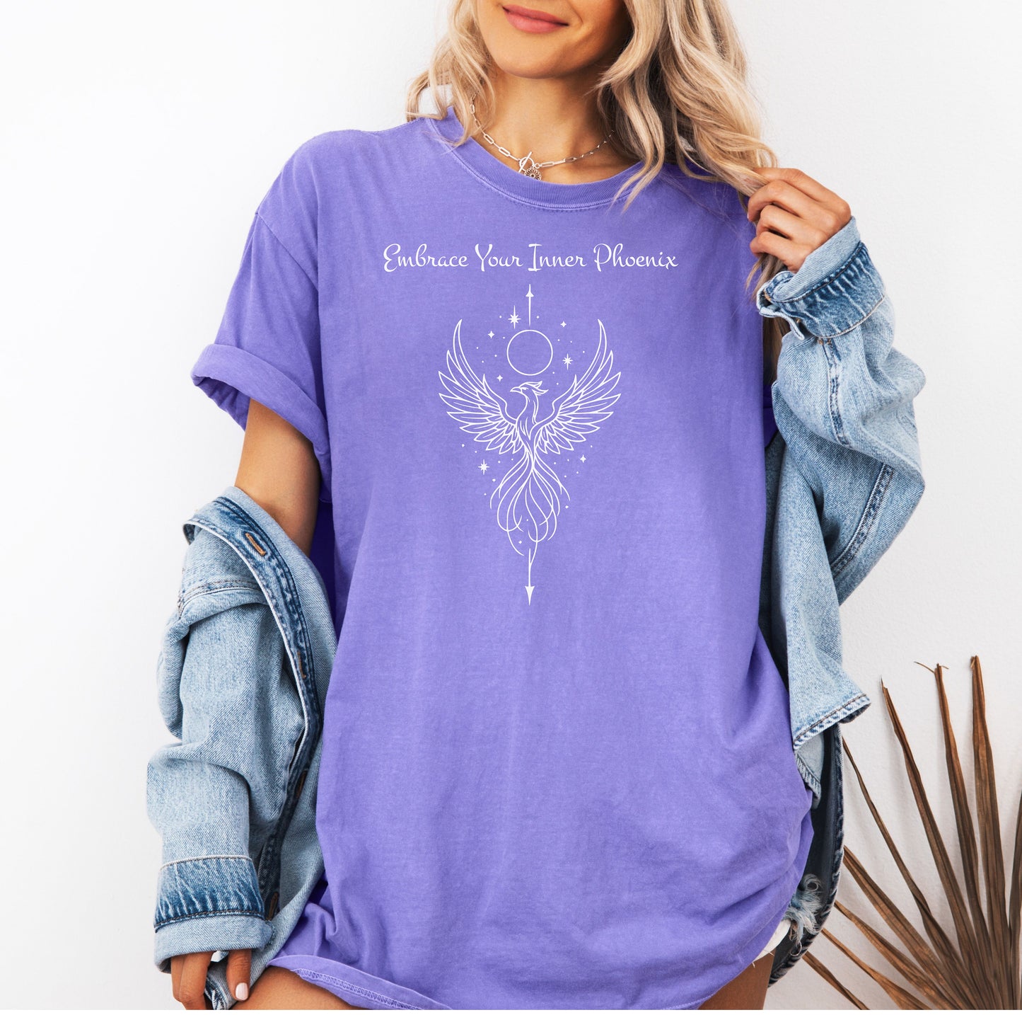 Person wearing a violet T-shirt featuring an elegant black line art phoenix design with wings spread wide, flowing tail feathers, and celestial elements including a moon and stars. Above the artwork, the phrase “Embrace Your Inner Phoenix” is printed in a graceful script font, symbolizing strength, rebirth, and transformation. Perfect for those who value empowerment, resilience, and a bold yet feminine style.