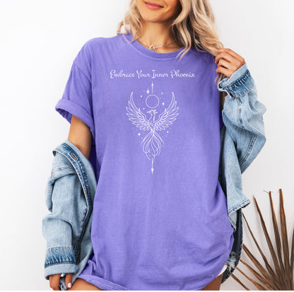 Person wearing a violet T-shirt featuring an elegant black line art phoenix design with wings spread wide, flowing tail feathers, and celestial elements including a moon and stars. Above the artwork, the phrase “Embrace Your Inner Phoenix” is printed in a graceful script font, symbolizing strength, rebirth, and transformation. Perfect for those who value empowerment, resilience, and a bold yet feminine style.