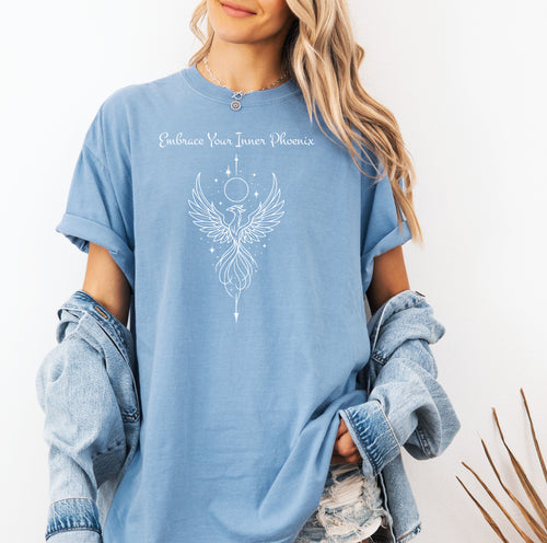 Person wearing a washed denim T-shirt featuring an elegant black line art phoenix design with wings spread wide, flowing tail feathers, and celestial elements including a moon and stars. Above the artwork, the phrase “Embrace Your Inner Phoenix” is printed in a graceful script font, symbolizing strength, rebirth, and transformation. Perfect for those who value empowerment, resilience, and a bold yet feminine style.