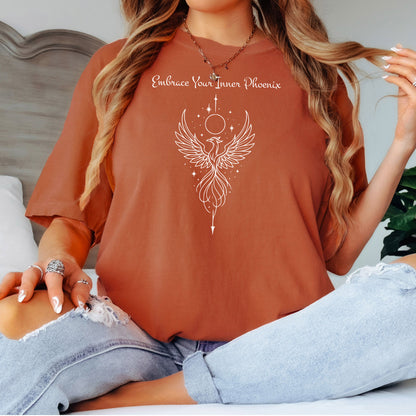 Person wearing a yam T-shirt featuring an elegant black line art phoenix design with wings spread wide, flowing tail feathers, and celestial elements including a moon and stars. Above the artwork, the phrase “Embrace Your Inner Phoenix” is printed in a graceful script font, symbolizing strength, rebirth, and transformation. Perfect for those who value empowerment, resilience, and a bold yet feminine style.