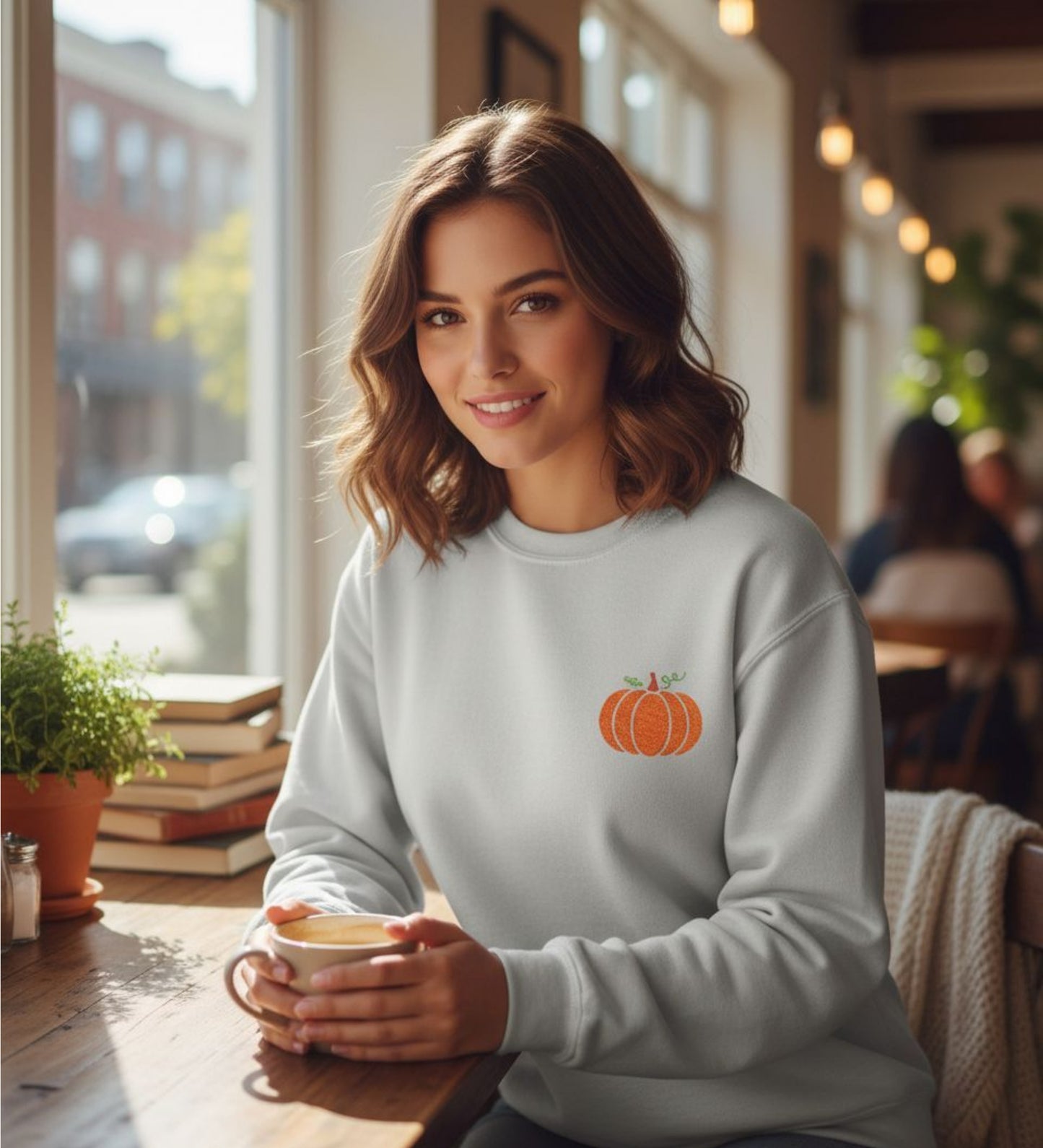 Person wearing an Ash sweatshirt with an embroidered pumpkin design, holding a coffee cup in a cozy cafe.