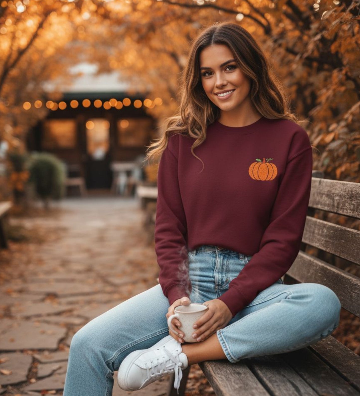 Person wearing a maroon sweatshirt with an embroidered pumpkin design, holding a coffee cup outdoors.