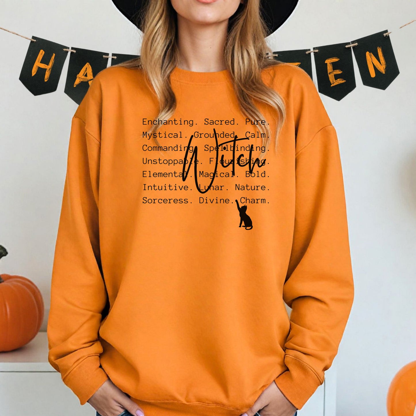 Person wearing a safety orange sweatshirt with Witch text and a black cat silhouette, standing in front of Halloween-themed decorations.