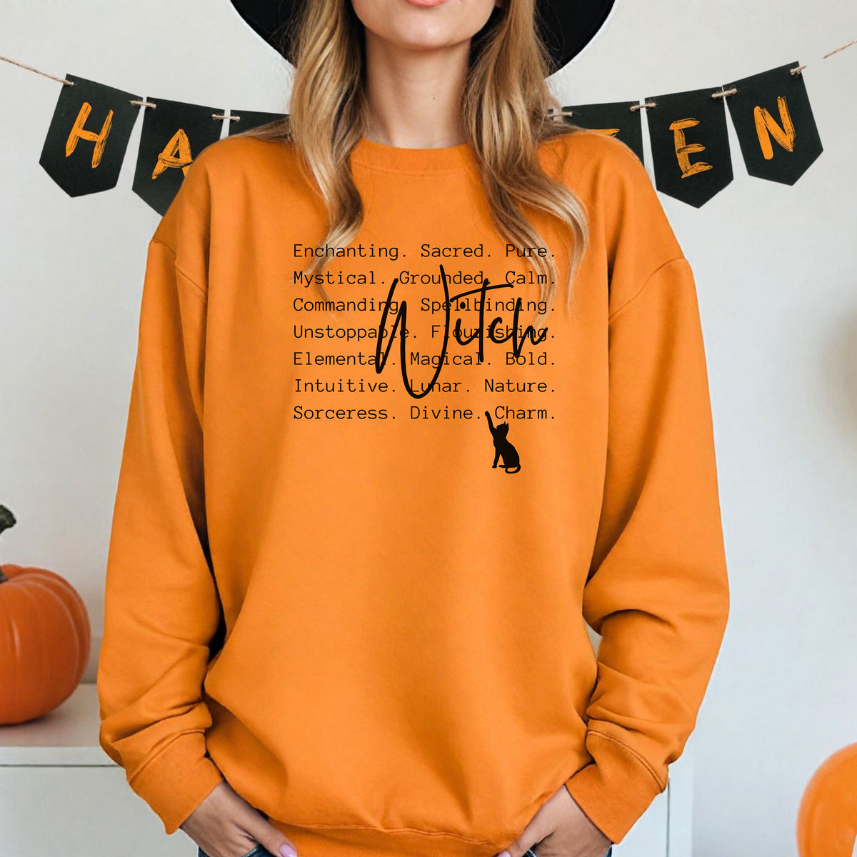 Person wearing a safety orange sweatshirt with Witch text and a black cat silhouette, standing in front of Halloween-themed decorations.