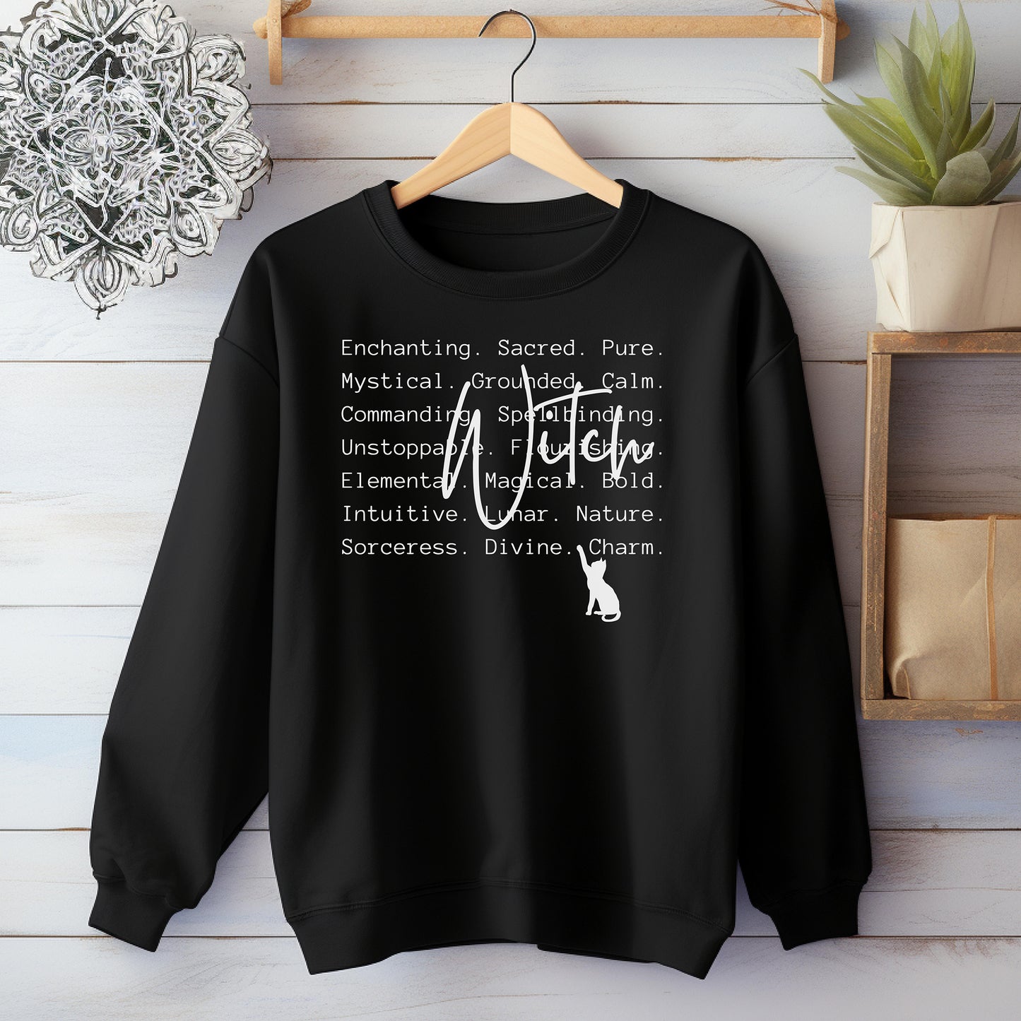 Black sweatshirt with Witch text and a cat graphic on a wooden hanger against a wooden wall.