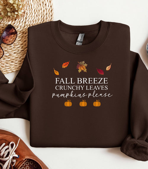 Dark Chocolate Gildan sweatshirt featuring a festive fall-themed design with colorful autumn leaves, the phrase “Fall Breeze, Crunchy Leaves, Pumpkins Please” in mixed fonts, and four orange pumpkin icons along the bottom. A cozy and stylish piece perfect for embracing crisp air, seasonal vibes, and pumpkin spice everything.
