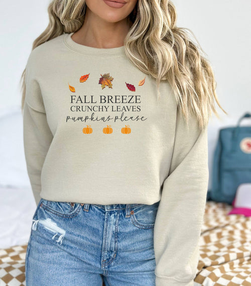 Sand Gildan sweatshirt featuring a festive fall-themed design with colorful autumn leaves, the phrase “Fall Breeze, Crunchy Leaves, Pumpkins Please” in mixed fonts, and four orange pumpkin icons along the bottom. A cozy and stylish piece perfect for embracing crisp air, seasonal vibes, and pumpkin spice everything.