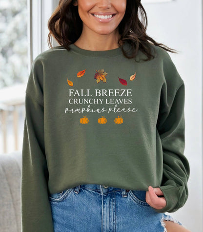 Military Green Gildan sweatshirt featuring a festive fall-themed design with colorful autumn leaves, the phrase “Fall Breeze, Crunchy Leaves, Pumpkins Please” in mixed fonts, and four orange pumpkin icons along the bottom. A cozy and stylish piece perfect for embracing crisp air, seasonal vibes, and pumpkin spice everything.