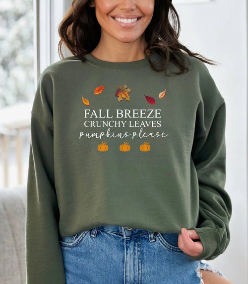 Military Green Gildan sweatshirt featuring a festive fall-themed design with colorful autumn leaves, the phrase “Fall Breeze, Crunchy Leaves, Pumpkins Please” in mixed fonts, and four orange pumpkin icons along the bottom. A cozy and stylish piece perfect for embracing crisp air, seasonal vibes, and pumpkin spice everything.