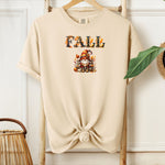 Ivory Comfort Colors T-shirt featuring a whimsical fall design with the word "FALL" in bold autumn-themed letters above a cozy gnome wearing a striped hat, surrounded by pumpkins, leaves, and acorns. The shirt is styled with a front knot and displayed on a wooden hanger in a rustic setting with natural accents.