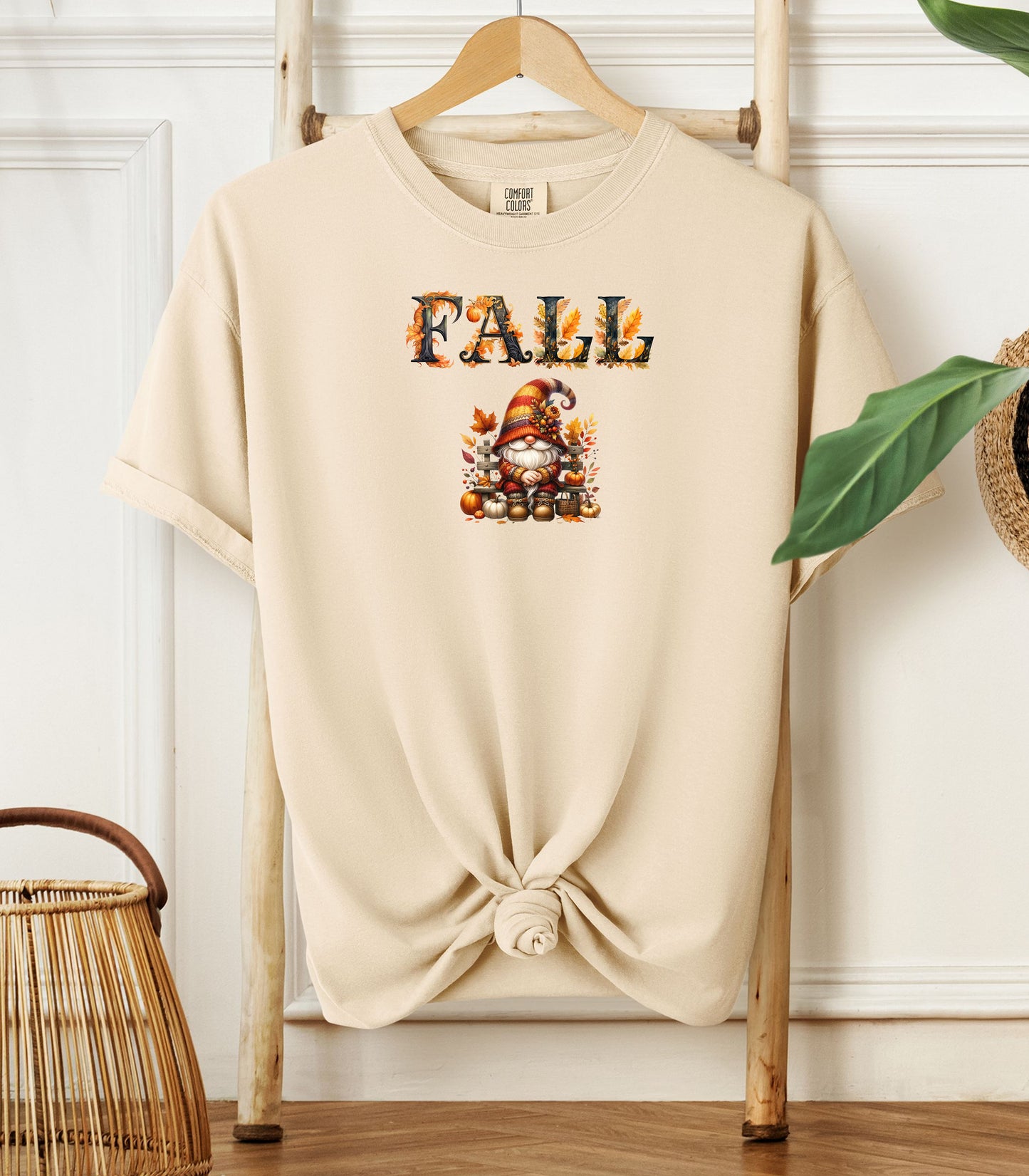 Ivory Comfort Colors T-shirt featuring a whimsical fall design with the word "FALL" in bold autumn-themed letters above a cozy gnome wearing a striped hat, surrounded by pumpkins, leaves, and acorns. The shirt is styled with a front knot and displayed on a wooden hanger in a rustic setting with natural accents.