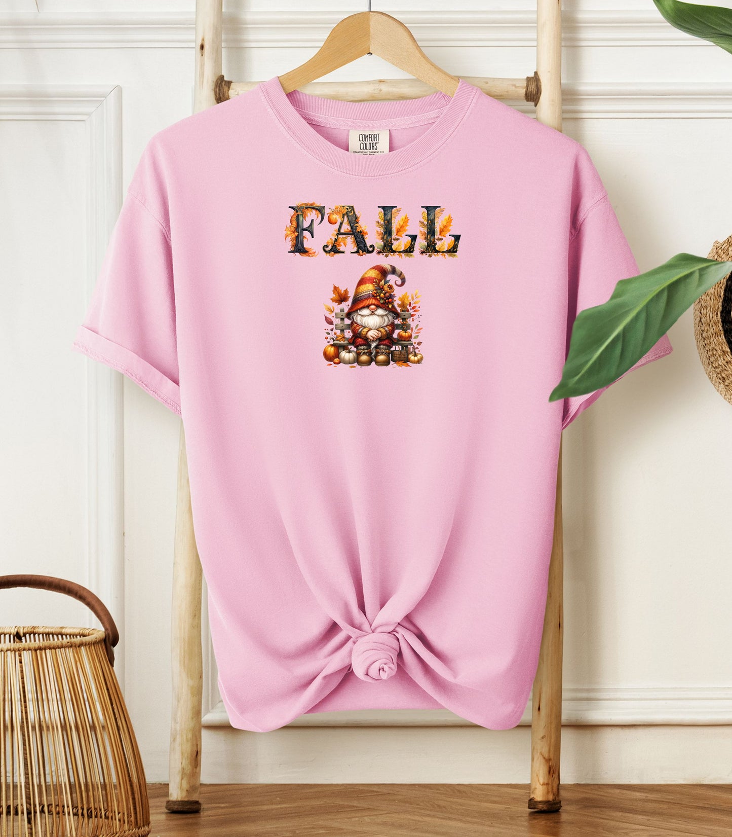 Blossom pink Comfort Colors T-shirt featuring a whimsical fall design with the word "FALL" in bold autumn-themed letters above a cozy gnome wearing a striped hat, surrounded by pumpkins, leaves, and acorns. The shirt is styled with a front knot and displayed on a wooden hanger in a rustic setting with natural accents.