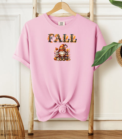 Blossom pink Comfort Colors T-shirt featuring a whimsical fall design with the word "FALL" in bold autumn-themed letters above a cozy gnome wearing a striped hat, surrounded by pumpkins, leaves, and acorns. The shirt is styled with a front knot and displayed on a wooden hanger in a rustic setting with natural accents.