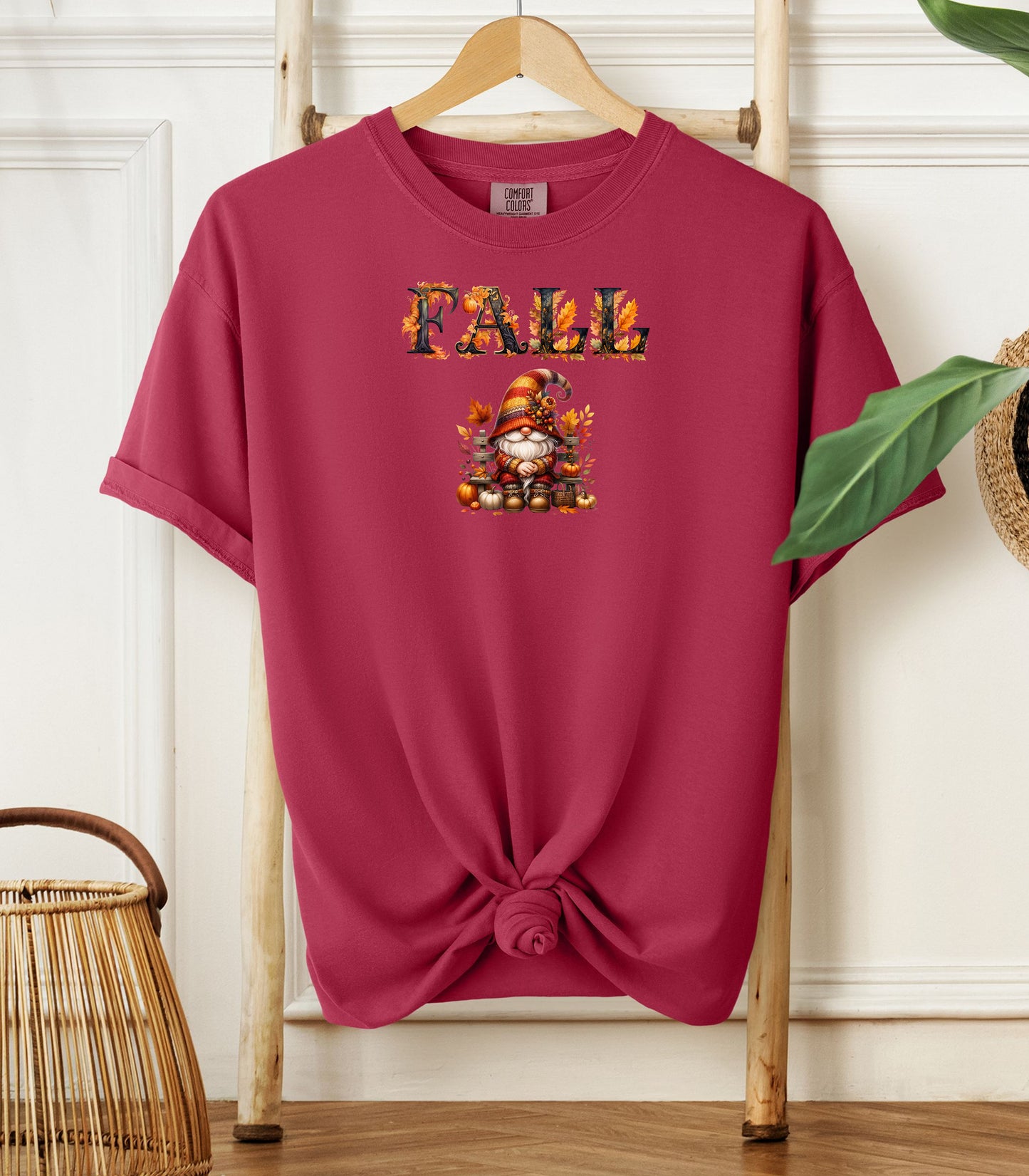 Chili Comfort Colors T-shirt featuring a whimsical fall design with the word "FALL" in bold autumn-themed letters above a cozy gnome wearing a striped hat, surrounded by pumpkins, leaves, and acorns. The shirt is styled with a front knot and displayed on a wooden hanger in a rustic setting with natural accents.