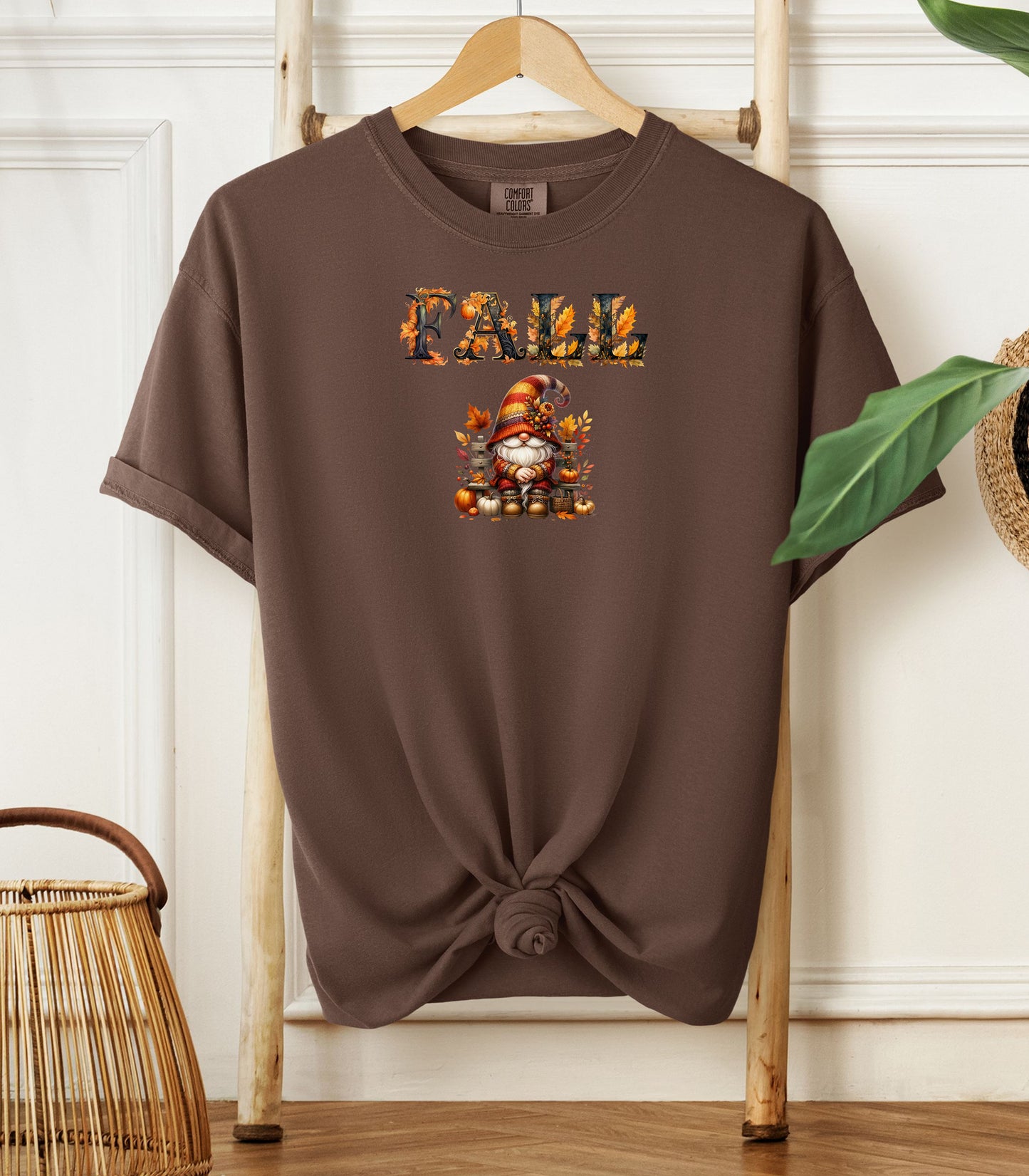 Espresso Comfort Colors T-shirt featuring a whimsical fall design with the word "FALL" in bold autumn-themed letters above a cozy gnome wearing a striped hat, surrounded by pumpkins, leaves, and acorns. The shirt is styled with a front knot and displayed on a wooden hanger in a rustic setting with natural accents.