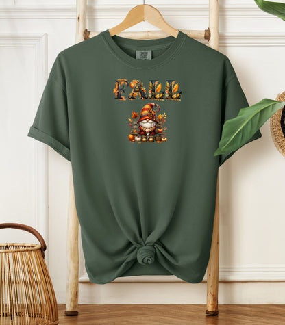 Moss Comfort Colors T-shirt featuring a whimsical fall design with the word "FALL" in bold autumn-themed letters above a cozy gnome wearing a striped hat, surrounded by pumpkins, leaves, and acorns. The shirt is styled with a front knot and displayed on a wooden hanger in a rustic setting with natural accents.