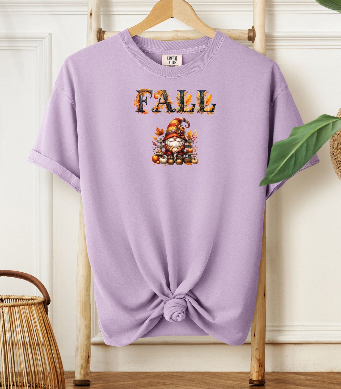 Orchid Comfort Colors T-shirt featuring a whimsical fall design with the word "FALL" in bold autumn-themed letters above a cozy gnome wearing a striped hat, surrounded by pumpkins, leaves, and acorns. The shirt is styled with a front knot and displayed on a wooden hanger in a rustic setting with natural accents.