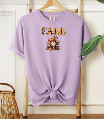 Orchid Comfort Colors T-shirt featuring a whimsical fall design with the word "FALL" in bold autumn-themed letters above a cozy gnome wearing a striped hat, surrounded by pumpkins, leaves, and acorns. The shirt is styled with a front knot and displayed on a wooden hanger in a rustic setting with natural accents.