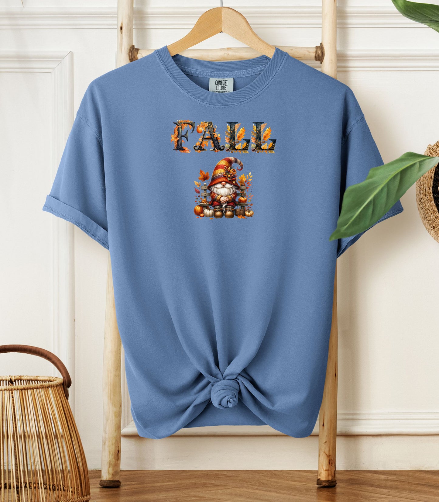 Washed Denim Comfort Colors T-shirt featuring a whimsical fall design with the word "FALL" in bold autumn-themed letters above a cozy gnome wearing a striped hat, surrounded by pumpkins, leaves, and acorns. The shirt is styled with a front knot and displayed on a wooden hanger in a rustic setting with natural accents.