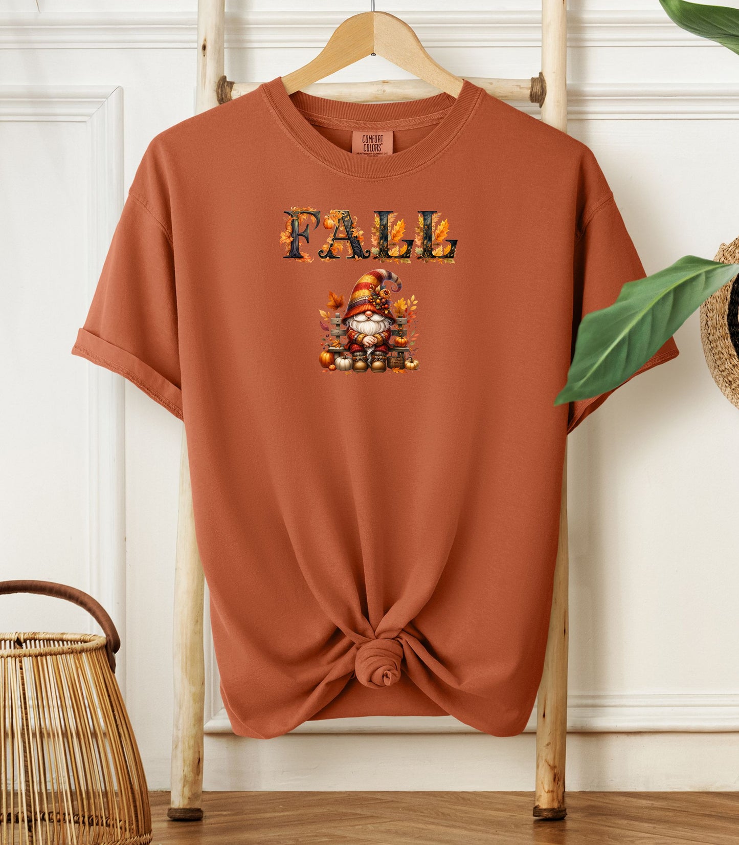 Yam Comfort Colors T-shirt featuring a whimsical fall design with the word "FALL" in bold autumn-themed letters above a cozy gnome wearing a striped hat, surrounded by pumpkins, leaves, and acorns. The shirt is styled with a front knot and displayed on a wooden hanger in a rustic setting with natural accents.