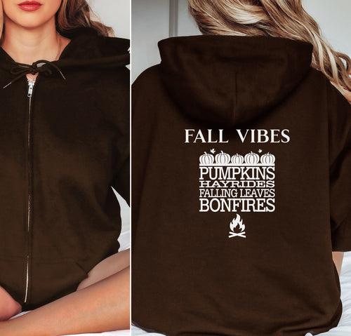 Dark Chocolate Gildan hoodie featuring a white“FALL VIBES” design on the back with stacked seasonal words: “PUMPKINS, HAYRIDES, FALLING LEAVES, BONFIRES,” framed by pumpkins above and a bonfire icon below. A cozy, festive graphic perfect for autumn lovers and fall adventures.