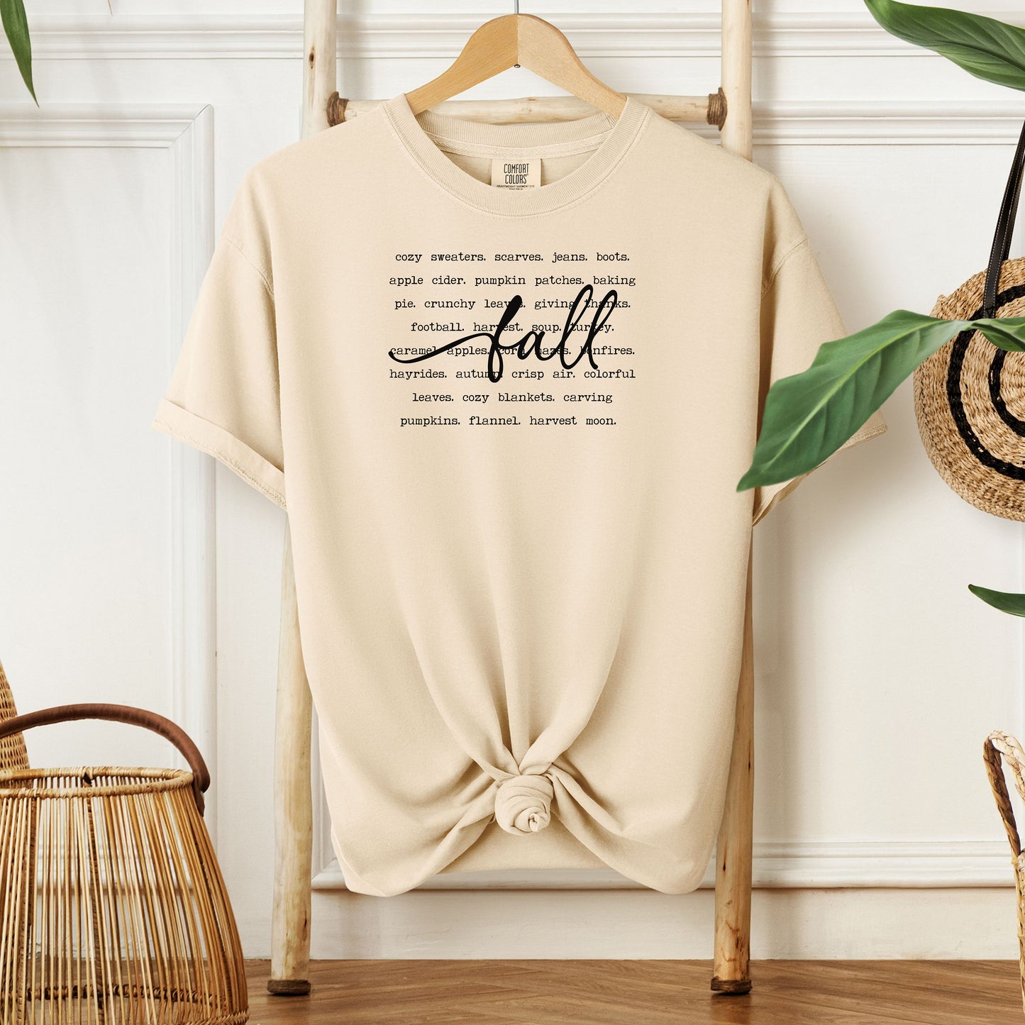 Ivory T-shirt with a cozy fall-themed text design featuring words like cozy sweaters, pumpkin patches, apple cider, hayrides, football, bonfires, flannel, and harvest moon. The word “Fall” stands out in a large handwritten script across the center, celebrating autumn vibes and seasonal favorites. Perfect for fall lovers and casual outfits.