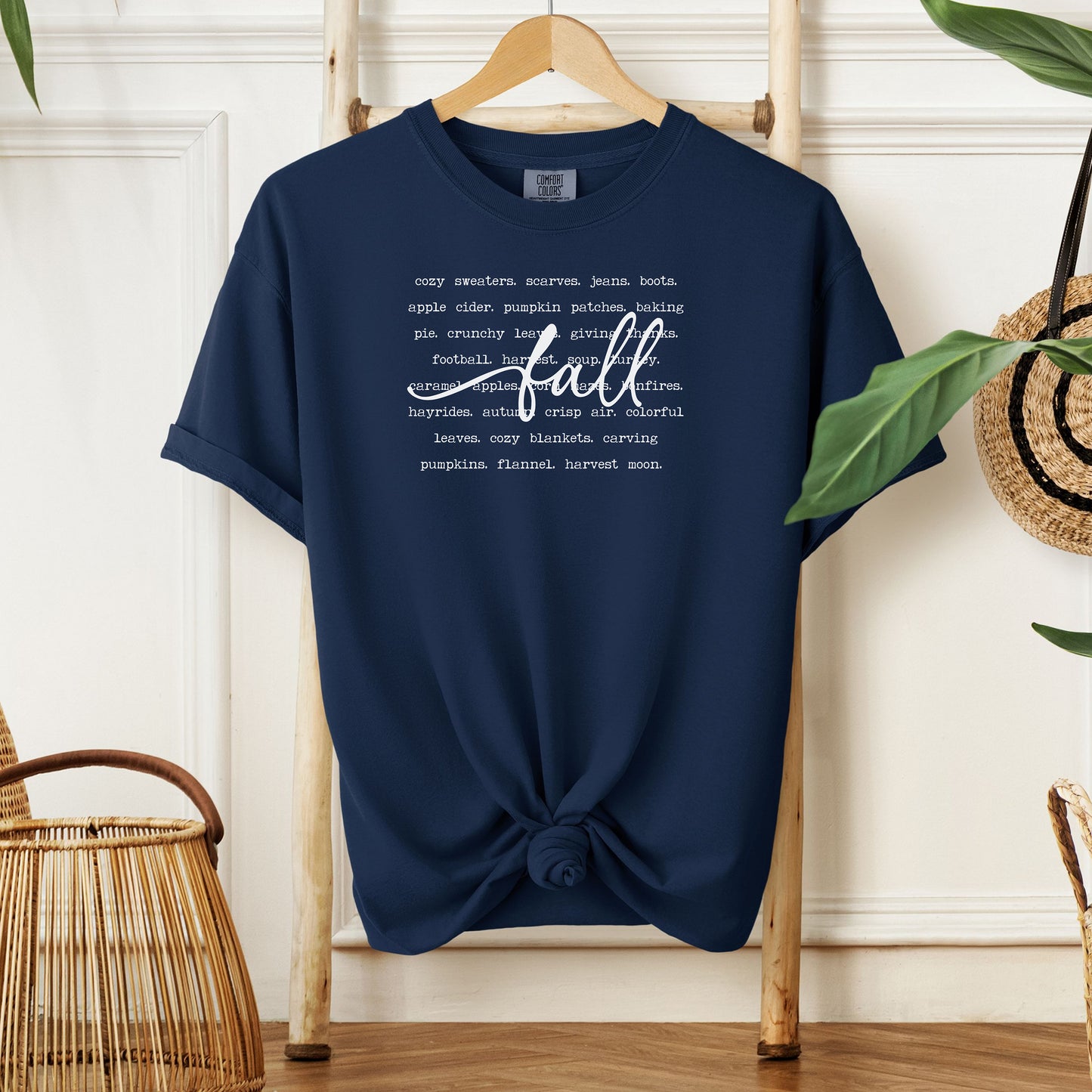True Navy T-shirt with a cozy fall-themed text design featuring words like cozy sweaters, pumpkin patches, apple cider, hayrides, football, bonfires, flannel, and harvest moon. The word “Fall” stands out in a large handwritten script across the center, celebrating autumn vibes and seasonal favorites. Perfect for fall lovers and casual outfits.