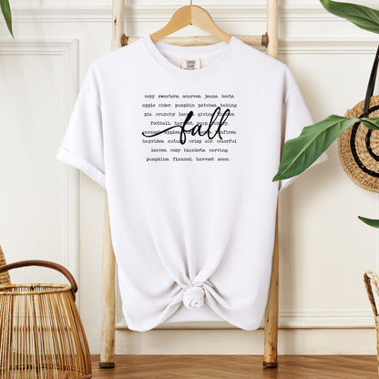White T-shirt with a cozy fall-themed text design featuring words like cozy sweaters, pumpkin patches, apple cider, hayrides, football, bonfires, flannel, and harvest moon. The word “Fall” stands out in a large handwritten script across the center, celebrating autumn vibes and seasonal favorites. Perfect for fall lovers and casual outfits.